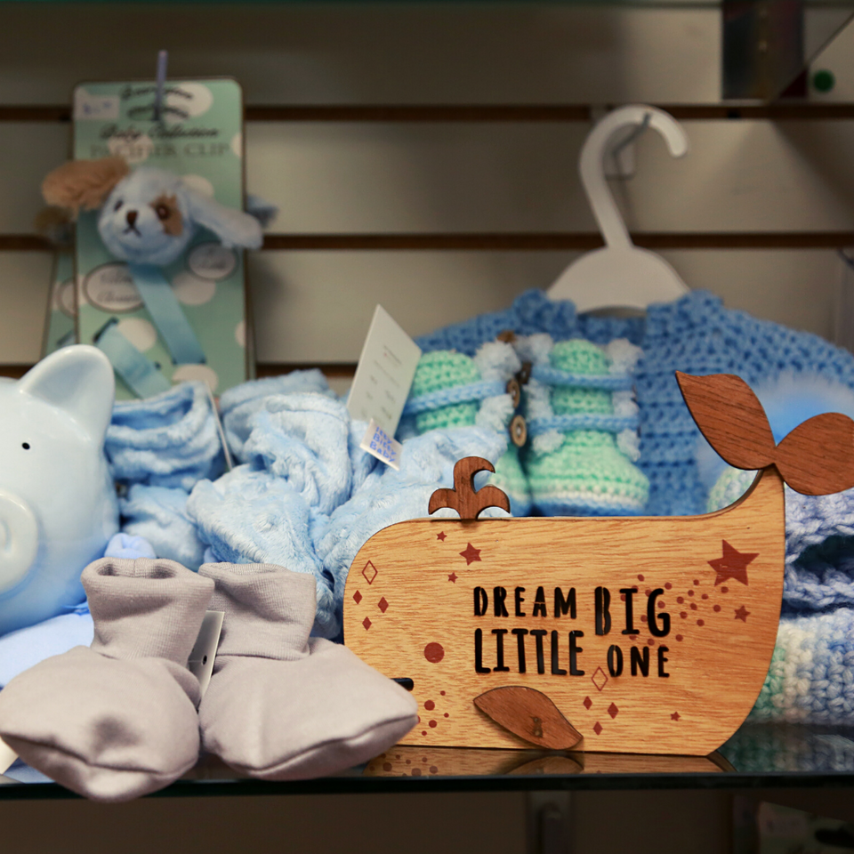 Shows a variety of different baby items that are for purchase. A cute whale that says, Dream Big Little One. A small pair of baby booties, a knitted sweater and a piggy bank. 