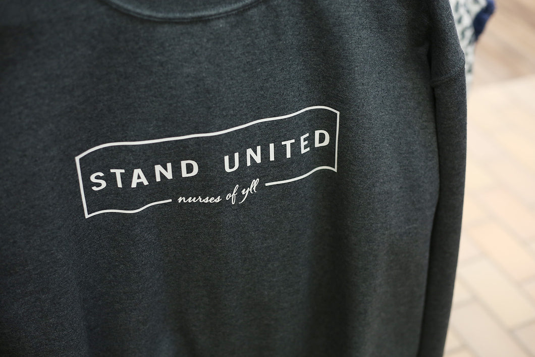 A dark grey sweater that has white text in the front that says, STAND UNITED nurses of YLL.